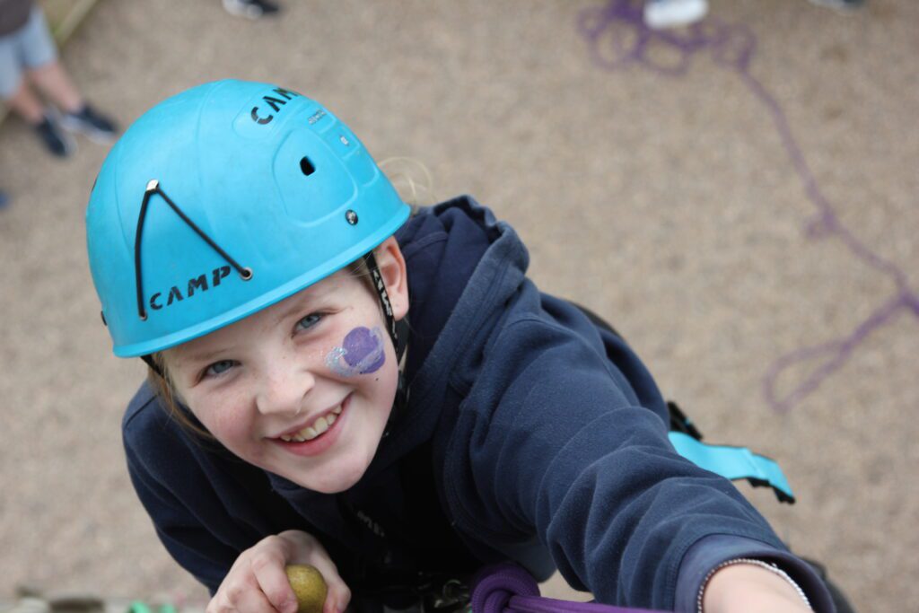 A view of Izzy on the climbing wall. The camera is at the top of the wall and Izzy is smiling up at it with a hard hat on and facepaint