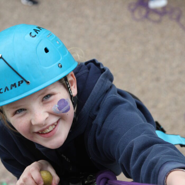 A view of Izzy on the climbing wall. The camera is at the top of the wall and Izzy is smiling up at it with a hard hat on and facepaint