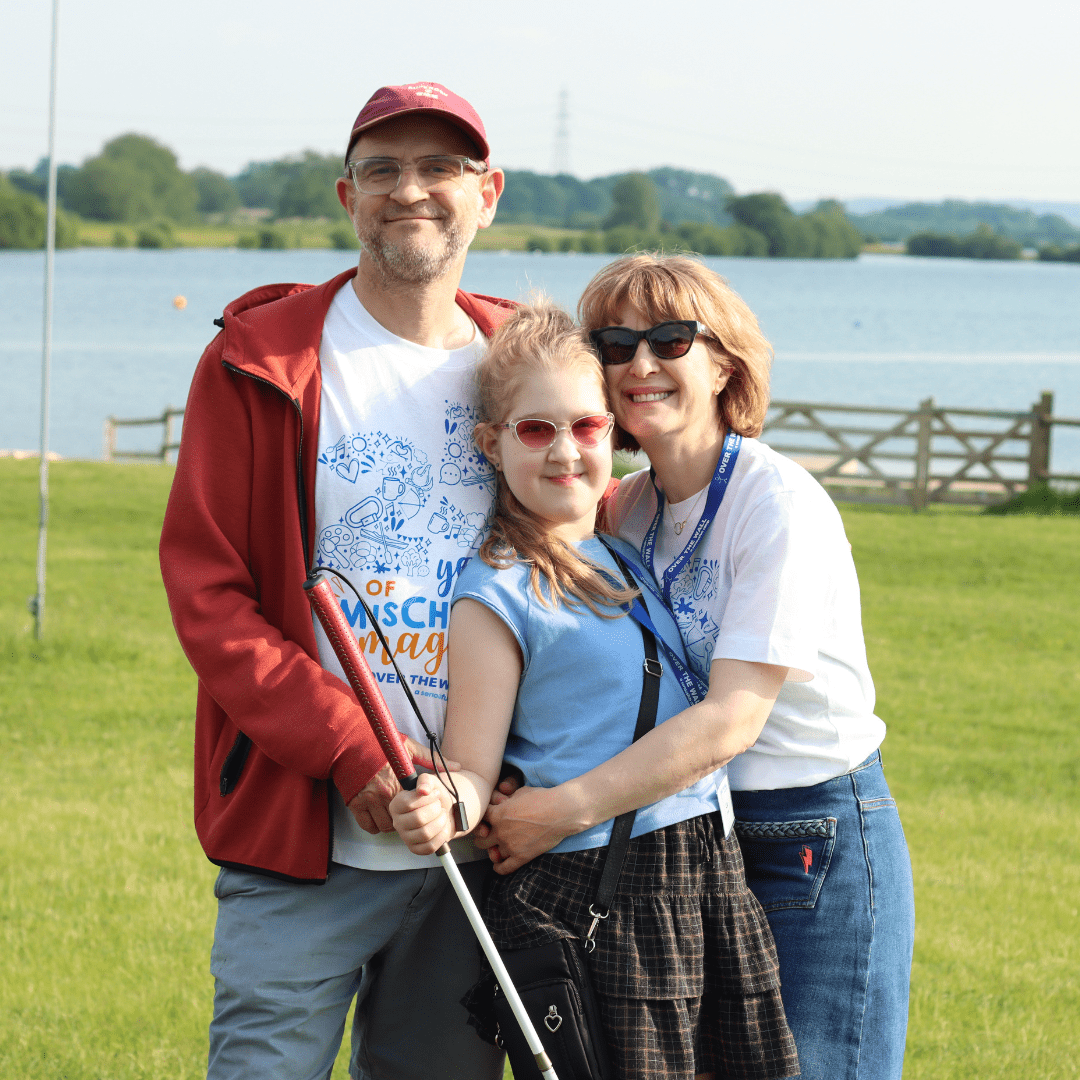 A family of three (mum, dad and daughter) are stood in front of a lake smiling at the camera with their arms around each other.