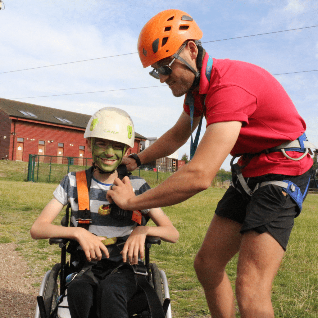 Young boy is sat in his wheelchair, having a harness fitted ready to take part in the climbing, abseiling and zipwire activities