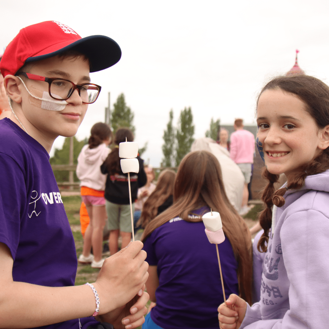 Young boy and girl stand opposite each other and smile at the camera. They are holding skewers with marshmallows on to roast on a fire.