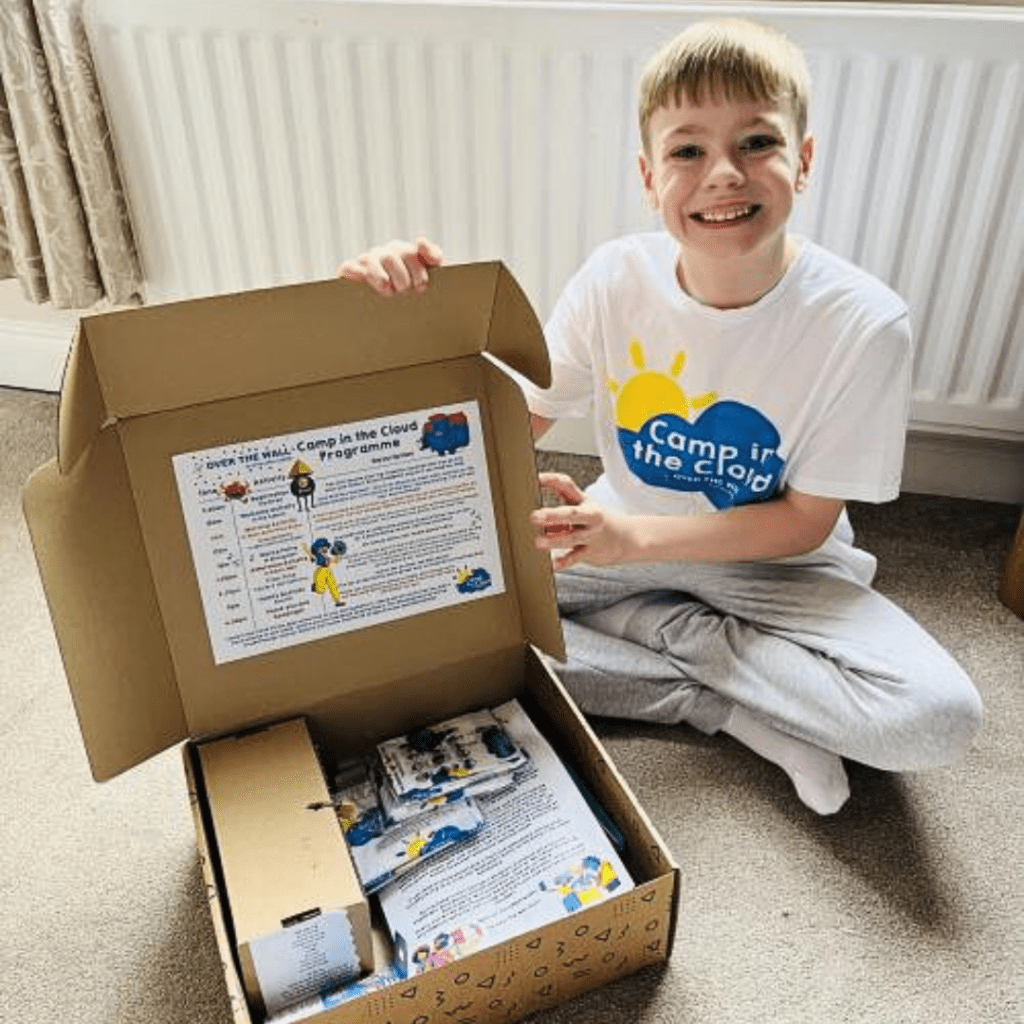 A boy is wearing a t-shirt with a Camp in the Cloud logo on it. He is smiling at the camera and showing his Seriously Fun Box