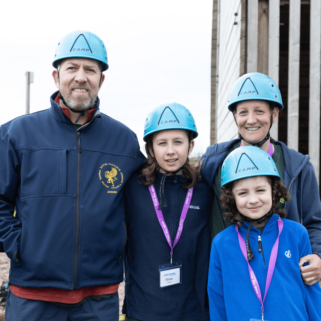 A family of four stand closely together by the climbing wall. They are wearing hard hats and all smiling at the camera.