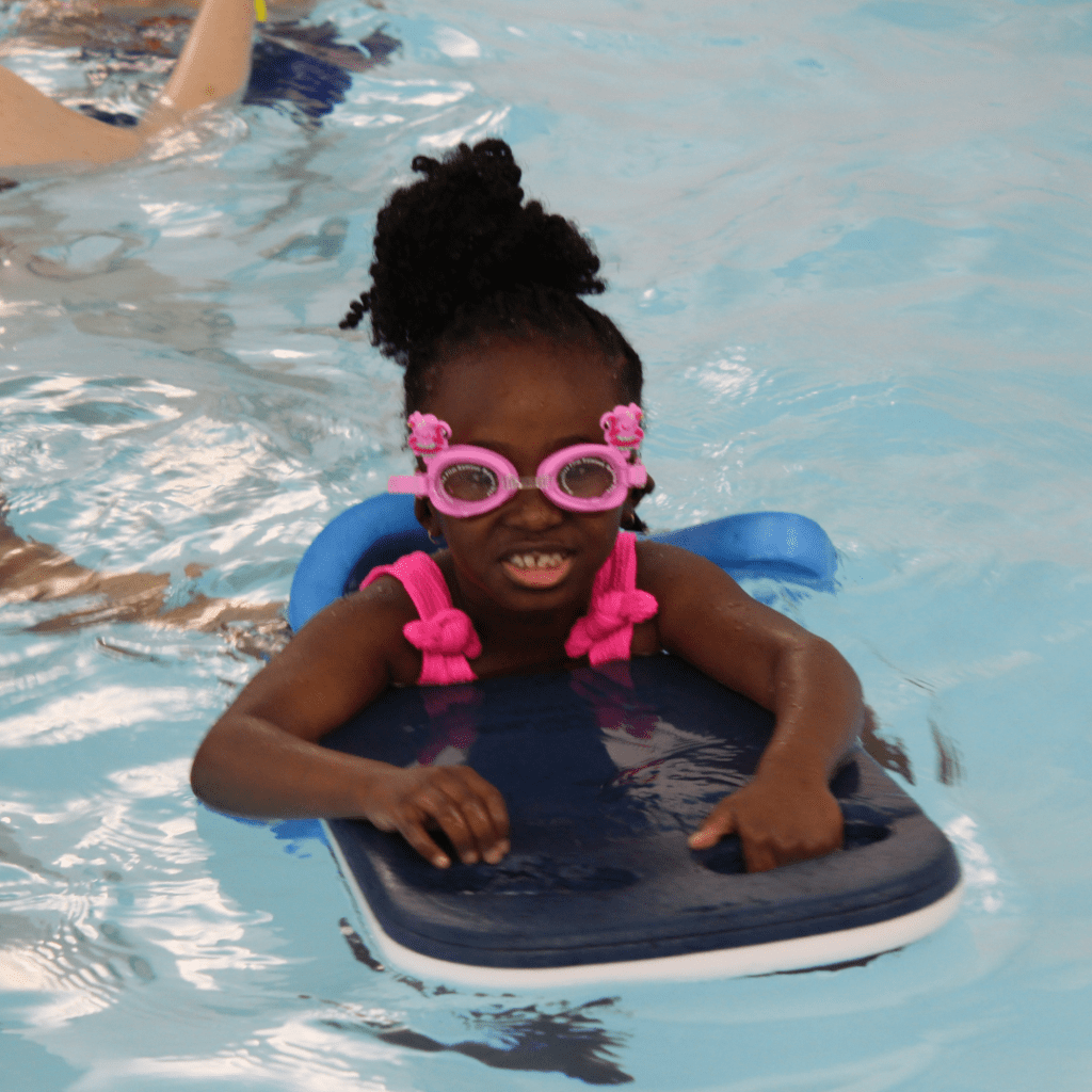 A girl in a swimming pool. She is wearing pink goggles and is holding onto a floating pad. She is smiling at the camera