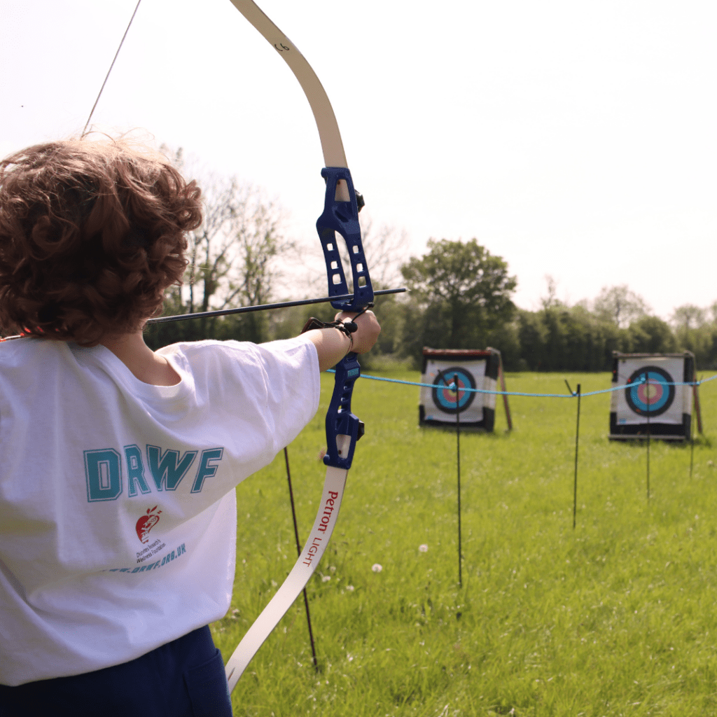 A child wearing a DRWF t-shirt is facing away from the camera and aiming a bow and arrow at an archery board.