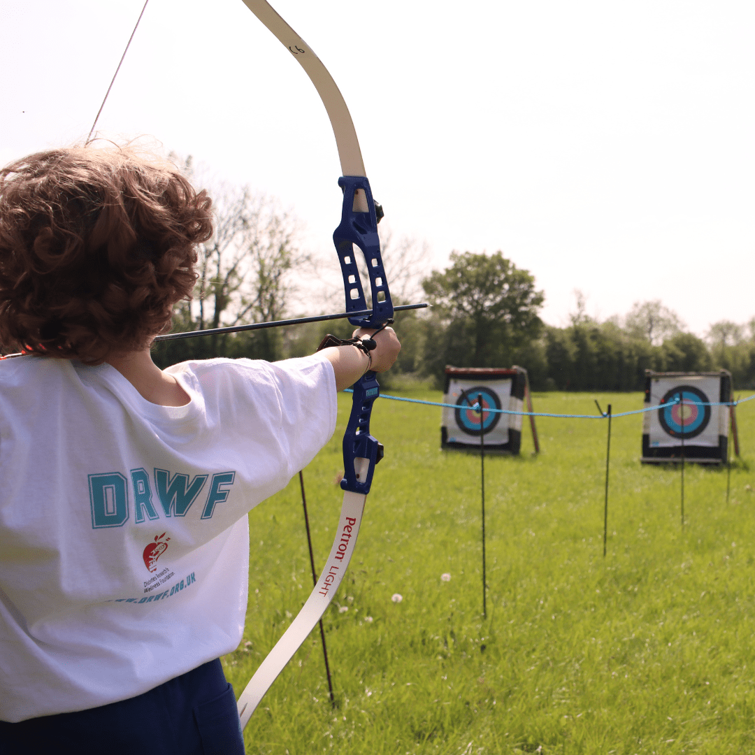 Magic (8) A child wearing a DRWF t-shirt is facing away from the camera and aiming a bow and arrow at an archery board.