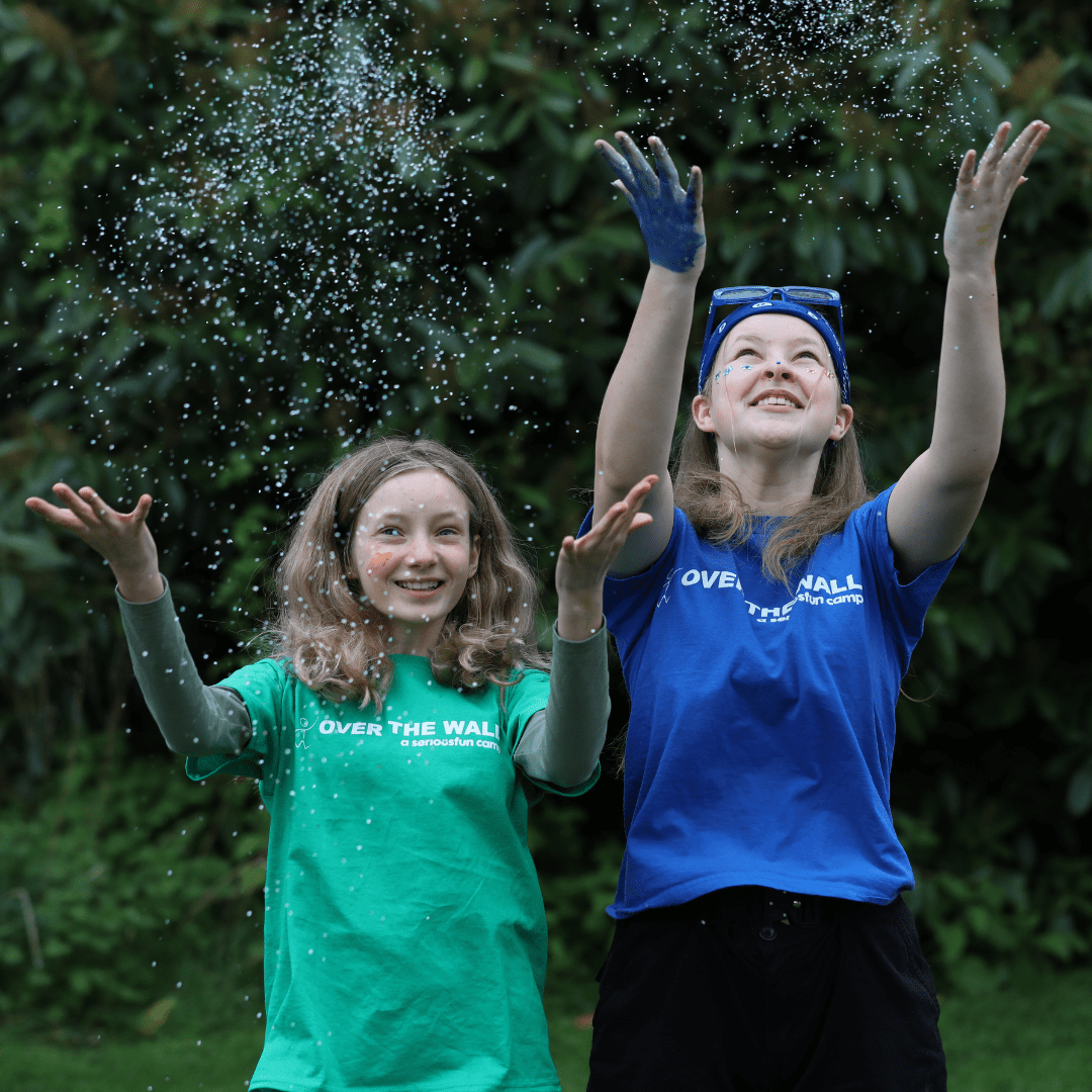 Two girls wearing Over The Wall Camp t-shirts smile and throw sparkles in the air