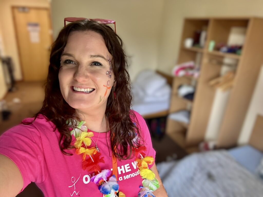 A selfie of Dr Zoe. She's smiling at the camera. She has a floral garland round her neck, pink heart-sharped glasses on her head, and face paint and glitter on.