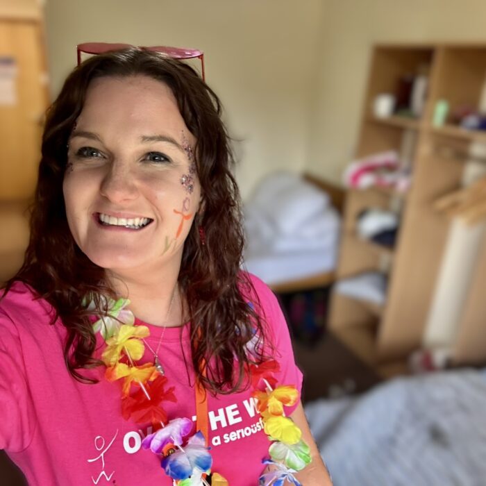 A selfie of Dr Zoe. She's smiling at the camera. She has a floral garland round her neck, pink heart-sharped glasses on her head, and face paint and glitter on.