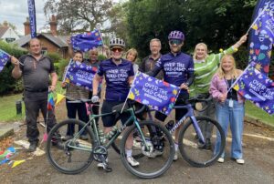 Group of Over The Wall Camp staff in t-shirts and waving flags with two cyclists at the front.