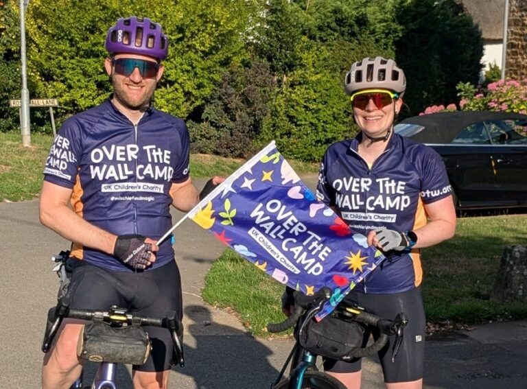 Man and woman wearing bike helmets and Over The Wall Camp cycling tops sitting on bikes and holding flags