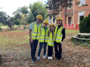 Lukass, Milana, and their parents are wearing hard hats and hi-vis vests, stood outside The Mount building where construction is starting. They are stood together smiling at the camera