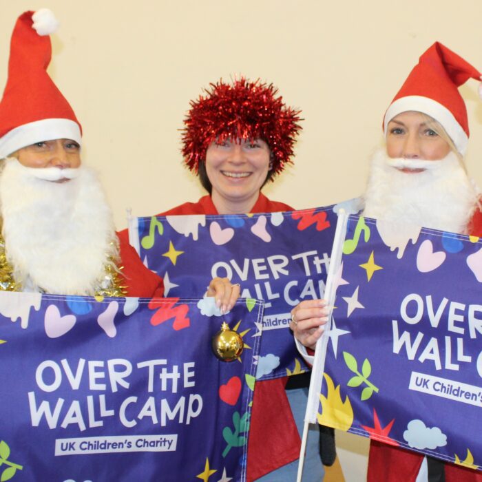 Three Over The Wall Camp staff dressed as Santa and with tinsel holding a flag