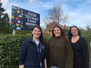 Three women with long dark hair smiling standing in front of a sign saying Over The Wall Camp