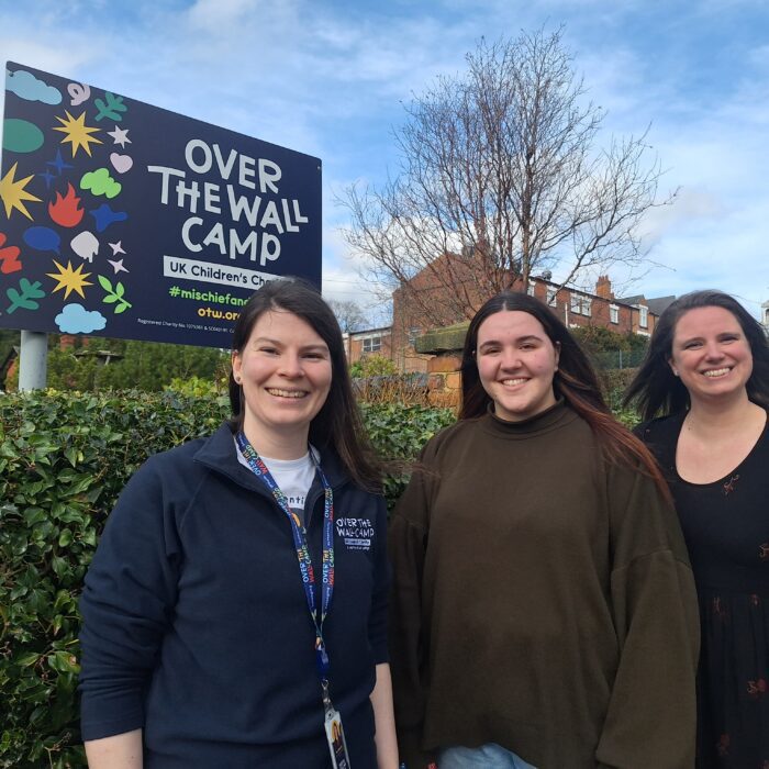Three women with long dark hair smiling standing in front of a sign saying Over The Wall Camp