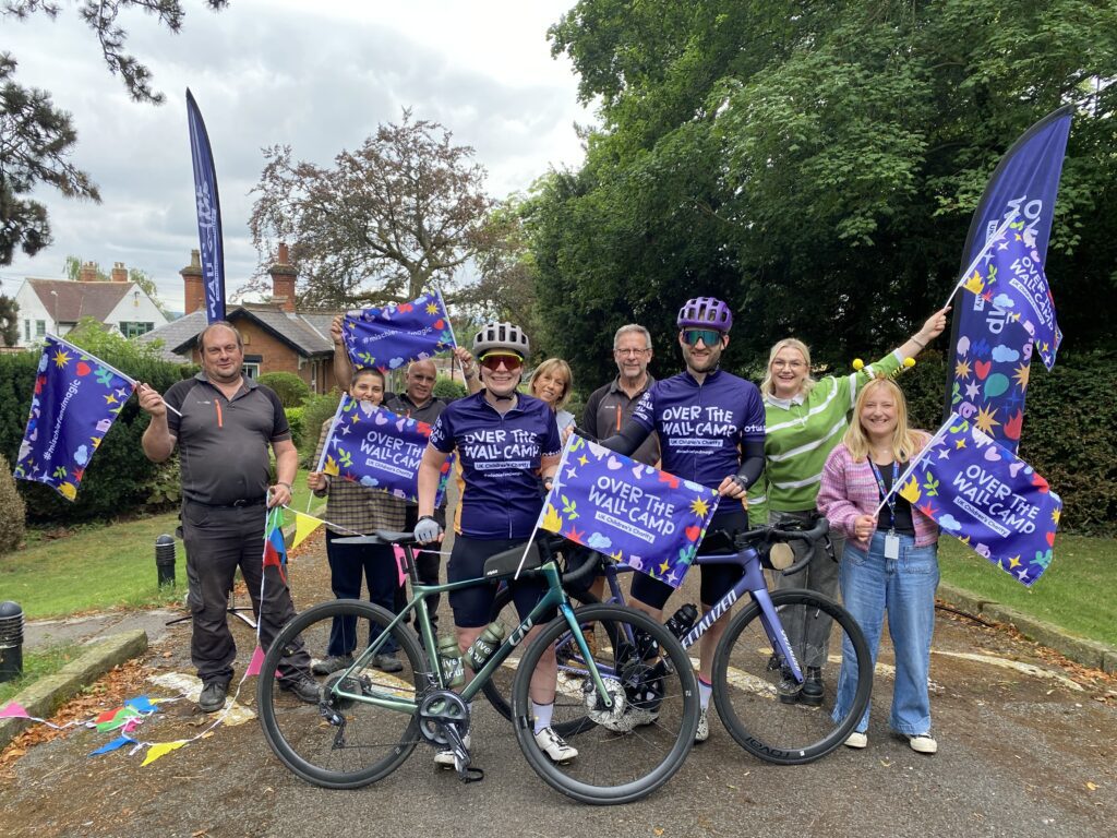 Group of people celebrating cyclists with Over The Wall Camp flags
