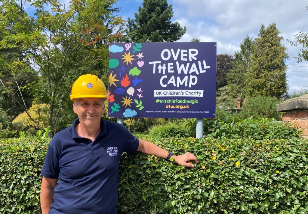Man in yellow hard hat and blue Over The Wall Camp shirt next to a sign saying Over The Wall Camp